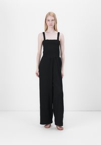 Mono negro con cuerpo fruncido, piernas anchas y tirantes gruesos. La tela tiene un aspecto texturizado, ideal para el uso casual.