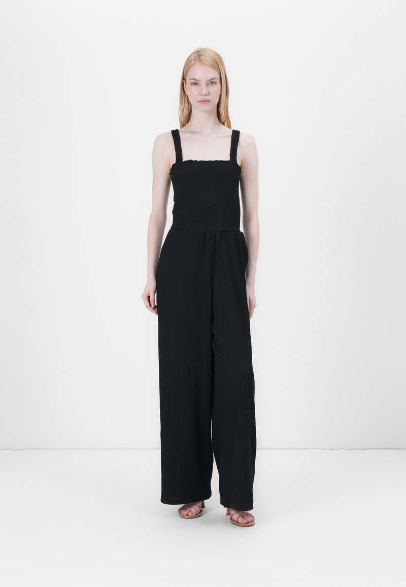 Mono negro con cuerpo fruncido, piernas anchas y tirantes gruesos. La tela tiene un aspecto texturizado, ideal para el uso casual.