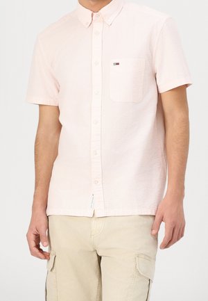 Mann trägt ein hellrosa kurzärmeliges Hemd mit Brusttasche und beige Cargoshorts, steht vor einem einfarbigen Hintergrund.