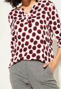 Blusa blanca con patrón espiral negro y rojo, mangas tres cuartos y escote en pico, combinada con pantalones grises. Textura de tela suave.