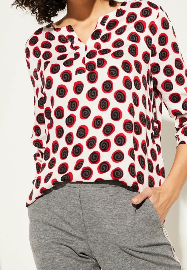Blusa blanca con patrón espiral negro y rojo, mangas tres cuartos y escote en pico, combinada con pantalones grises. Textura de tela suave.