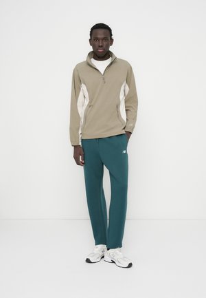 Polo zippé kaki avec des accents blancs, associé à un pantalon de survêtement bleu sarcelle foncé. Le modèle porte des baskets blanches et grises. Coupe simple et sportive.