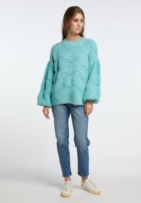 Un pullover spesso di colore blu chiaro con un motivo a onde testurizzato, caratterizzato da maniche ampie e una vestibilità rilassata, abbinato a jeans chiari e sneakers.