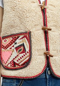 Μπεζ γιλέκο από φλις με υφή, με τσέπη με μοτίβο σε κόκκινο, ροζ και μαύρο, και ξύλινες κλειδαριές-κουμπιά.