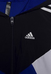 Giacca sportiva nera e blu con chiusura a zip, fodera in rete e logo Adidas ricamato sul davanti. Accenti grigi vicino all'orlo.