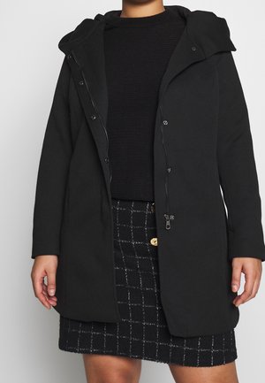 Frau trägt einen schwarzen Kapuzenmantel über einem schwarzen Pullover und einem schwarzen Rock mit weißem Fenstergittermuster und Holzknöpfen.