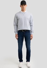 Młody mężczyzna w czarnej czapce, szarej bluzie na białej koszulce, ciemnoniebieskich dżinsach i białych tenisówkach stoi na jednolitym jasnoszarym tle.