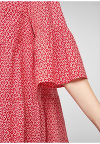 Manche de robe rouge avec motif circulaire géométrique blanc, coupe ample et poignet légèrement évasé, montrant un bras partiel sur fond gris.