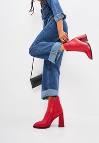 Bottes en cuir rouge montantes avec talon épais, fermeture éclair latérale et bout arrondi, portées avec un jean denim retroussé. Sac noir minimaliste visible.