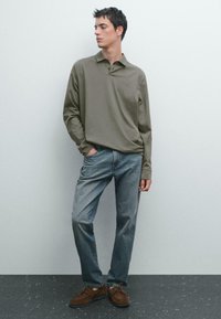 Olijfgroene polo met lange mouwen en een kraag, gecombineerd met lichte blauwe spijkerbroek en bruine suède schoenen. Casual pasvorm, minimale details.