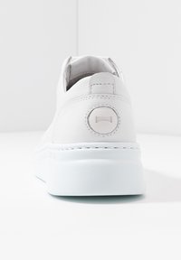 Chaussure blanche en cuir à enfiler avec un accent rond à l'arrière, semelle texturée et design minimaliste. Détails de couture visibles.