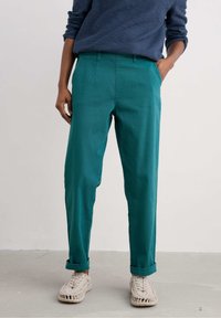 Pantalons teal ajustés avec des plis avant et des ourlets roulés, fabriqués à partir d'un tissu texturé. Associés à des chaussures tissées de couleur claire.