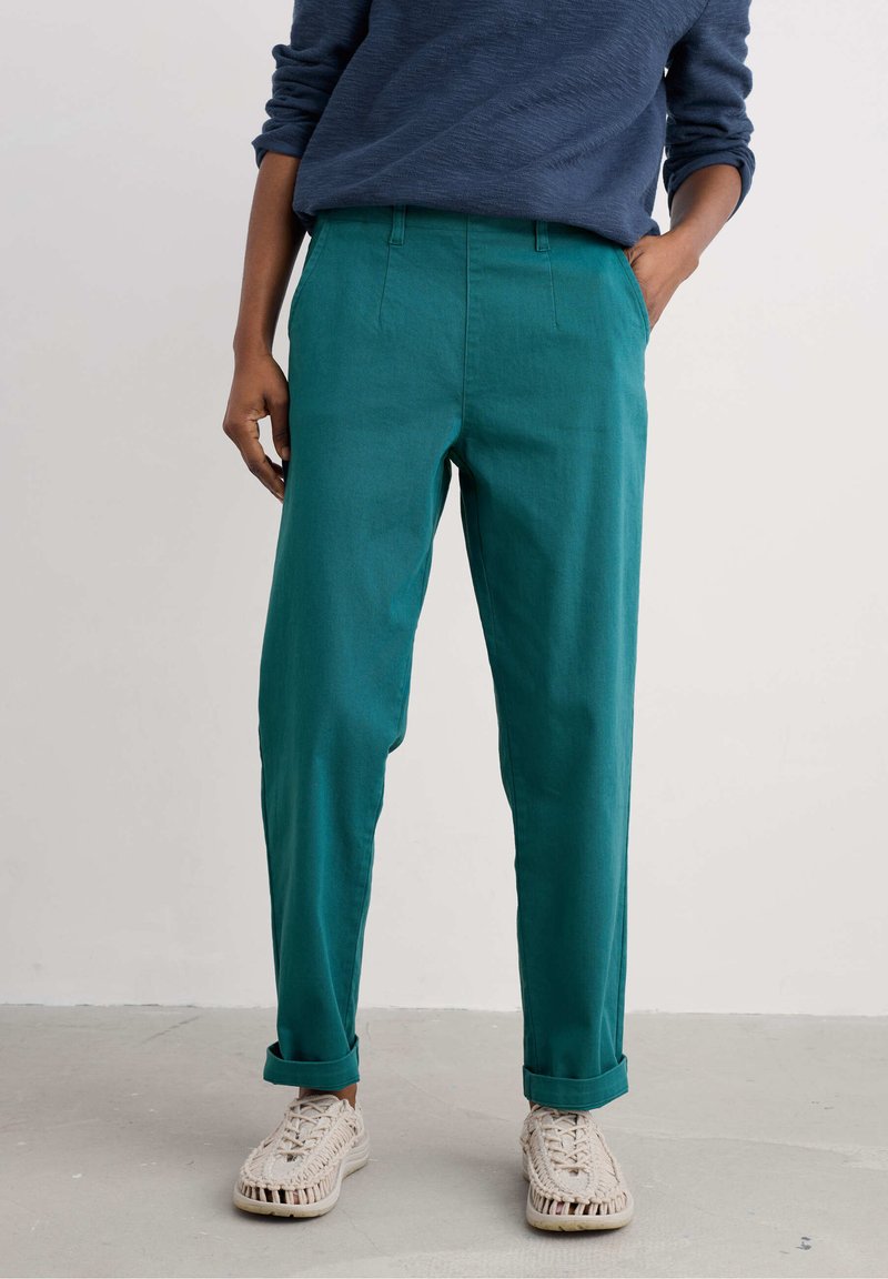 Pantalons teal ajustés avec des plis avant et des ourlets roulés, fabriqués à partir d'un tissu texturé. Associés à des chaussures tissées de couleur claire.