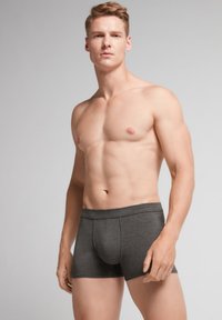 Graue Boxer-Shorts aus weichem Material, mit einer engen Passform, elastischem Bund und einer Öffnung vorne. Das Modell steht in neutraler Pose.