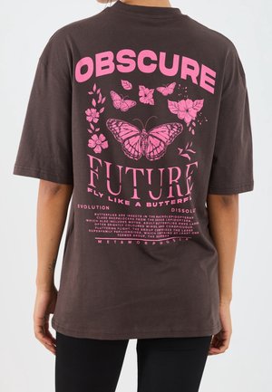 Persoon die een donkerbruine T-shirt draagt met roze vlinder- en bloemafbeeldingen en de tekst "OBSCURE FUTURE Fly like a butterfly" op de rug.