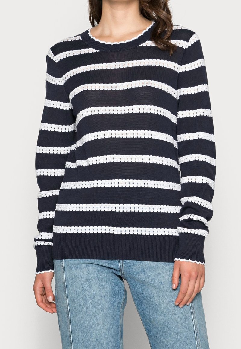 Femme portant un pull en maille bleu marine à rayures horizontales blanches et bords festonnés, associé à un jean denim bleu clair.