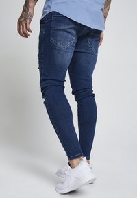 Vaqueros ajustados de mezclilla azul con detalles desgastados, diseño ceñido, cintura de mediana altura y dos bolsillos traseros. Combinados con zapatillas deportivas blancas.