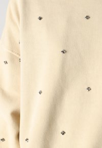 Sweatshirt beige clair avec une texture douce, comportant de petites embellissements floraux en argent et des accents foncés répartis uniformément.