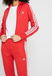 Červený adidas teplákový komplet obsahující zipovou bundu s bílými pruhy na rukávech a odpovídající kalhoty, oboje s logovými akcenty a hladkou texturou.