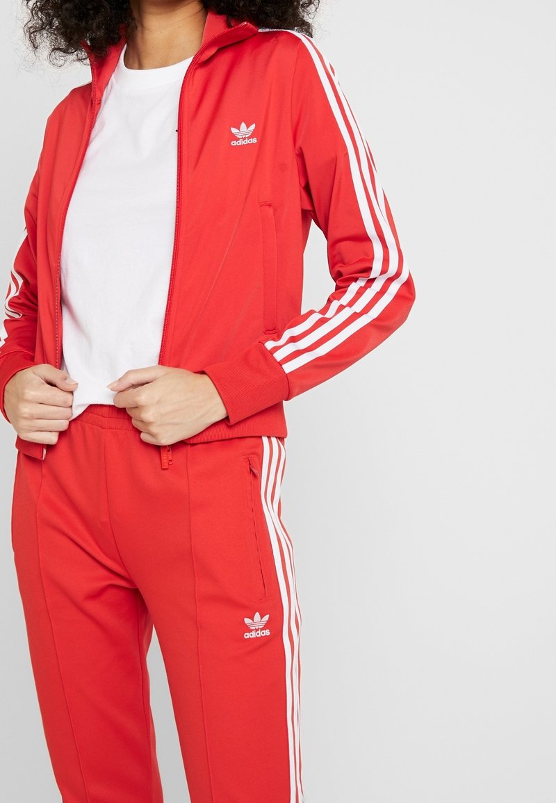 Červený adidas teplákový komplet obsahující zipovou bundu s bílými pruhy na rukávech a odpovídající kalhoty, oboje s logovými akcenty a hladkou texturou.
