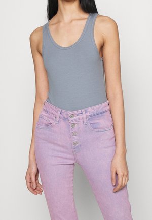 Femme portant un débardeur gris à côtes sans manches rentré dans un jean rose taille haute avec fermeture à boutons, se tenant devant un fond uni.