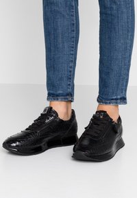 Černé tenisky s texturou hadí kůže a elegantním designem. Mají černé šněrování, zipový detail a lesklou podrážku.