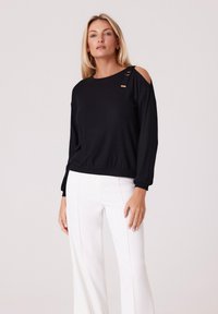 Czarny top z długim rękawem w fasonie typu cold shoulder, z złotymi akcentami i prążkowaną teksturą, zestawiony z białymi spodniami o szerokich nogawkach.