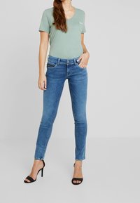 Maglietta verde a scollo a V con dettaglio tasca, abbinata a jeans skinny blu. L'outfit è completato da sandali neri con tacco alto e cinturini.