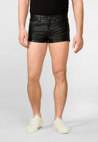 Zwarte leren shorts met een nauwsluitend ontwerp, voorzien van een voorknopsluiting, riemlussen en zijzakken, gecombineerd met crèmekleurige sneakers.