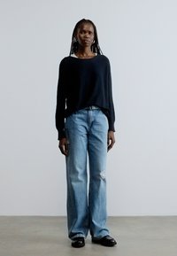 Musta neulottu neulepaita, vaaleansiniset kulutetut leveälahkeiset farkut ja mustat kengät. Malli seisoo yksinkertaisen vaalean taustan edessä.