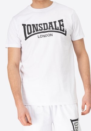 Man draagt een wit Lonsdale London T-shirt en bijpassende witte shorts met het Lonsdale-logo op het been, staand tegen een effen achtergrond.
