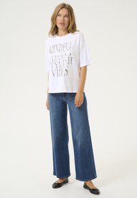 T-shirt en coton blanc avec un texte métallisé argent affichant "VIVRE EN CONSCIENCE, VIBES STYLÉES", associé à un pantalon en denim bleu à jambes larges et des ballerines noires.