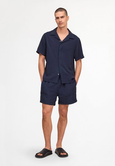 Homme aux cheveux courts debout, portant une chemise à manches courtes bleu marine boutonnée, un short assorti et des sandales noires à enfiler. Mains dans les poches.