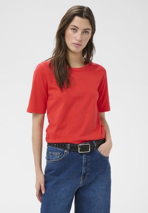 Mujer joven con cabello largo y castaño, vistiendo una camiseta roja de manga corta, jeans azules y un cinturón tejido negro, de pie frente a un fondo liso.