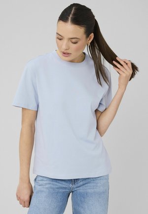 Femme aux longs cheveux foncés attachés en queue de cheval, portant un t-shirt bleu clair à manches courtes et un jean bleu, sur un fond clair uni.