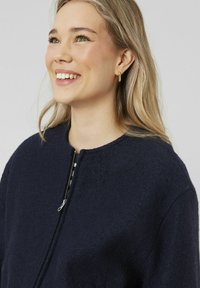 Veste texturée bleu marine avec un col rond et une fermeture éclair à l'avant, fabriquée dans un matériau doux semblable à de la laine et avec des détails de design minimalistes.