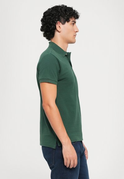 Camisa polo verde hecha de tejido texturizado, con cuello, mangas cortas y un dobladillo recto. Combinada con jeans azul oscuro.