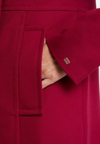 Abrigo de textura roja con un bolsillo lateral y un puño con un acento plateado. La tela parece duradera y estructurada.