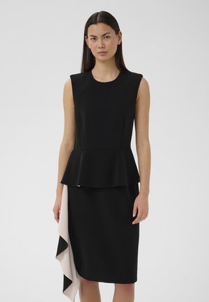 Žena s rovnými tmavými vlasmi, oblečená v čiernych šatách bez rukávov s volánom v páse a asymetrickým bielo-čiernym zavinovacím detailom na sukni.