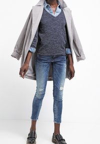 Abrigo gris sobre un suéter de pico azul marino y una camisa de mezclilla azul clara, combinado con jeans skinny azules desgastados y zapatos planos negros.