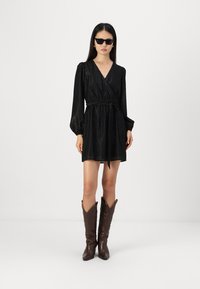 Fekete, hosszú ujjú, texturált anyagú wrap dress és meghúzott derék. Barna cowboy csizmával és sötét napszemüveggel párosítva.