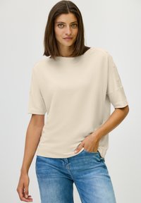 Beige short-sleeve top gemaakt van zachte stof, met een relaxte pasvorm, ronde hals en decoratieve knopen op de schouders.
