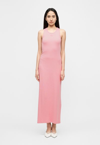 Ροζ ριμπ μακρύ φόρεμα χωρίς μανίκια, με εφαρμοστή σιλουέτα και διακριτική λεπτομέρεια με λογότυπο στο ντεκολτέ. Το μοντέλο φοράει λευκά μυτερά παπούτσια.