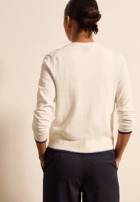 Osoba z ciemnymi włosami w kok jedzie białą, długoskalową bluzkę i ciemne spodnie, stojąc z rękami w kieszeniach przy beżowej ścianie.