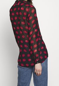 Gennemsigtig sort bluse med levende røde lippetryk, knap-ned stil, lange ærmer og en afslappet pasform, parret med lyseblå denim jeans.