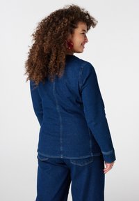 Spijkerjack met lange mouwen, diepblauwe kleur, contraststiksels, getailleerd model, zichtbare naden, gedragen met bijpassende spijkerbroek.