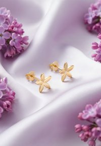 Boucles d'oreilles en forme de fleur en or reposant sur un tissu satiné lavande, entourées de fleurs de lilas violettes. Texture lisse avec une finition poli.