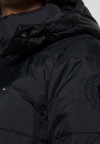 Sort puffers jakke med hætte, lavet af glinsende nylonmateriale, med quiltet struktur og et rundt logo på ærmet.