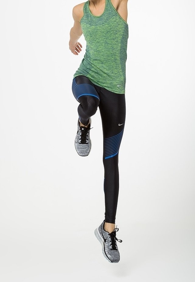 Personne portant un haut sans manches vert et des leggings noirs à rayures bleues, sautant ou courant sur fond blanc.