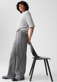 Grijze corduroy wijde broek en een verlopen lichtgrijs gebreid trui. Het model staat naast een moderne zwarte stoel met strakke lijnen.